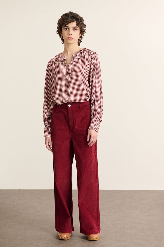 Mannequin femme de face portant une blouse en vichy bordeaux, col grand à volant, coupe droite et fermeture boutonnée.