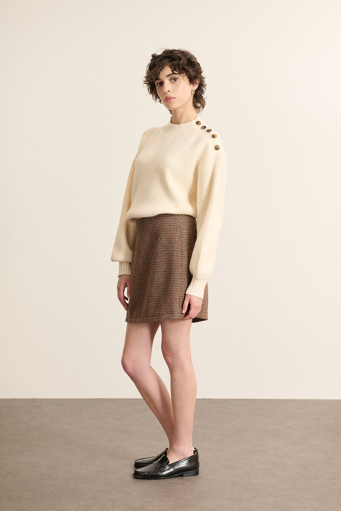 Mannequin femme de face portant un pull ample écru, col rond, manches longues et ouverture boutonnée sur l’épaule gauche.